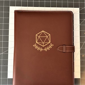 May include: A brown leather folder with a gold-colored D20 dice symbol and a strap closure.