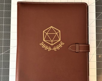 May include: A brown leather folder with a gold-colored D20 dice symbol and a strap closure.