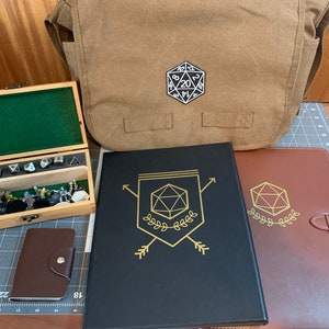 May include: A brown leather messenger bag with a white 20-sided die patch.  A wooden box with dice and miniature figures. Two notebooks with gold foil designs of a 20-sided die.
