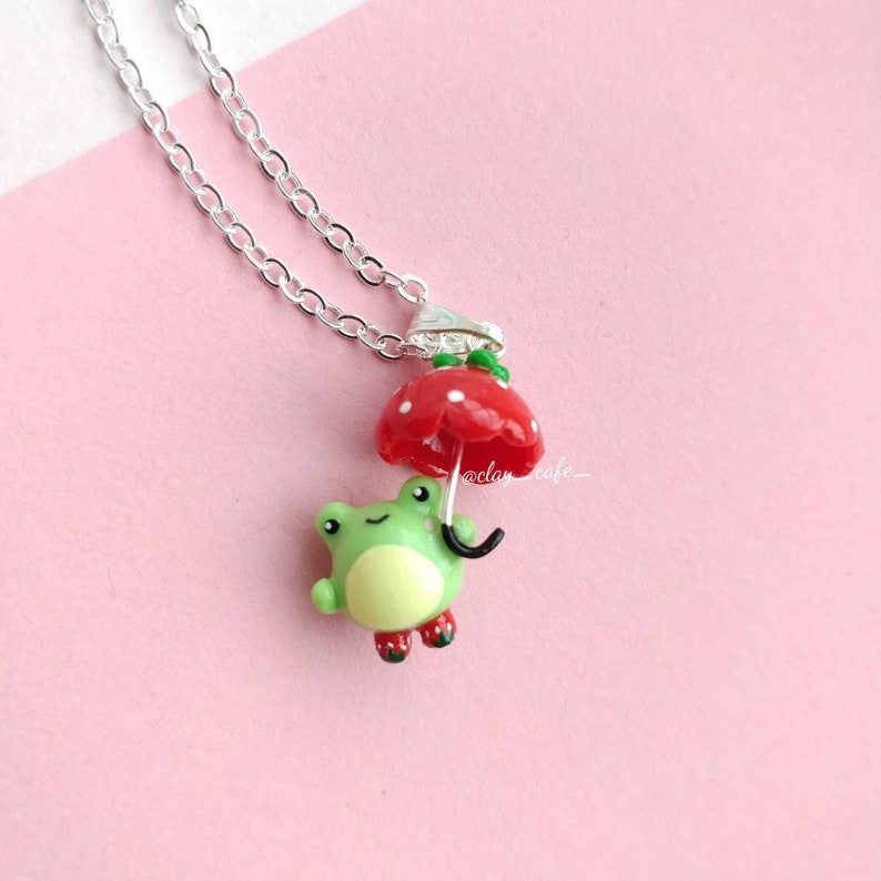 Pu&ograve; includere: Una collana con una rana verde con stivali rossi che tiene un ombrello rosso con pois bianchi. La collana &egrave; fatta di una catena d'argento.