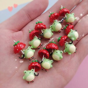 Pu&ograve; includere: Una collezione di ciondoli in pasta polimerica fatti a mano con rane verdi che tengono ombrelli rossi a forma di fragola. I ciondoli sono perfetti per aggiungere un tocco fantasioso a gioielli, portachiavi o altri oggetti artigianali.