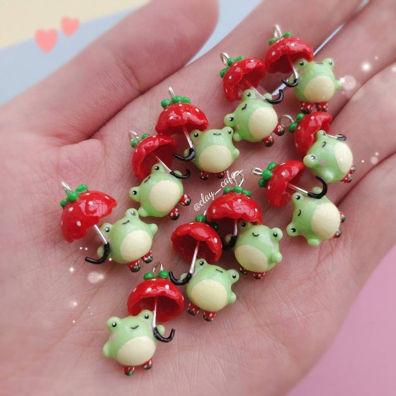 Pu&ograve; includere: Una collezione di ciondoli in pasta polimerica fatti a mano con rane verdi che tengono ombrelli rossi a forma di fragola. I ciondoli sono perfetti per aggiungere un tocco fantasioso a gioielli, portachiavi o altri oggetti artigianali.