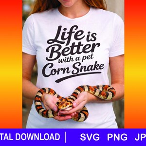 May include: A white t-shirt with the text "Life is Better with a pet Corn Snake" in black. A corn snake with orange, black, and white markings is held in front of the shirt. The image has a gradient orange and yellow background.
