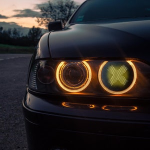 Puede incluir: Un coche negro con las luces delanteras encendidas. Los faros son un anillo de luz amarilla alrededor de una luz blanca más pequeña y brillante. El coche está aparcado en una carretera oscura.