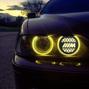 Może przedstawiać: Zbliżenie na ciemnobrązowy samochód z żółtymi reflektorami. Reflektory mają wzór plastra miodu, a logo BMW jest widoczne w środku prawego reflektora.