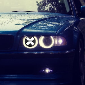 May include: A blue BMW sedan featuring a front grille with the BMW emblem and dual circular headlights. The driver-side headlight displays a bright white circular halo ring with a central black and white X-shaped graphic featuring the M logo.