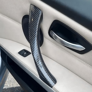 Puede incluir: Primer plano del interior de una puerta de coche. El panel de la puerta es de color crema con un tirador negro y plateado. El tirador tiene un patrón de fibra de carbono. La puerta también tiene un reposabrazos negro y un pequeño altavoz.