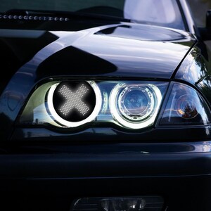 May include: A black car with a white X in the headlight. The car is parked on a paved road with green grass in the background.