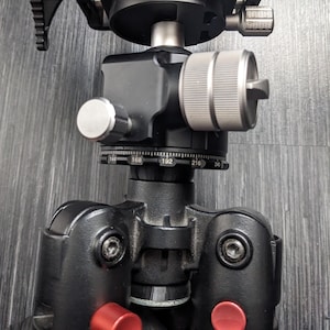 May include: A black tripod with a silver and black camera mount. The tripod has three legs and a center column. The camera mount has a knob for adjusting the angle of the camera. The camera mount has a scale with numbers from 144 to 240.