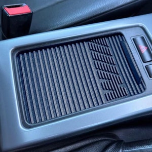 May include: A close-up of a car's interior, showing a gray and black textured panel with a small red warning light. The panel has a series of vertical lines and a small rectangular opening.