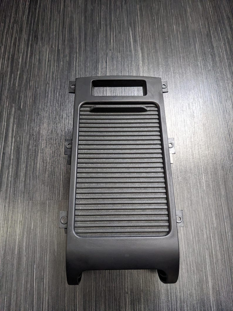 May include: Black plastic car part with a rectangular opening and a series of horizontal lines. The part has a curved bottom and several small holes on the sides.
