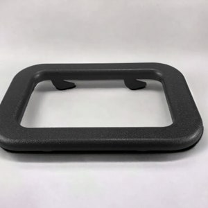 Puede incluir: Un marco de pl&aacute;stico negro con dos peque&ntilde;as pesta&ntilde;as en la parte superior. El marco est&aacute; dise&ntilde;ado para sujetar una placa de matr&iacute;cula.
