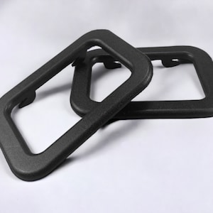 Puede incluir: Dos marcos rectangulares de pl&aacute;stico negro con bordes redondeados. Los marcos tienen una superficie texturizada y una peque&ntilde;a abertura en un lado.