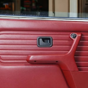 Puede incluir: El interior de una puerta de coche roja con una manija de puerta negra y un reposabrazos rojo. El panel de la puerta tiene una textura acanalada.