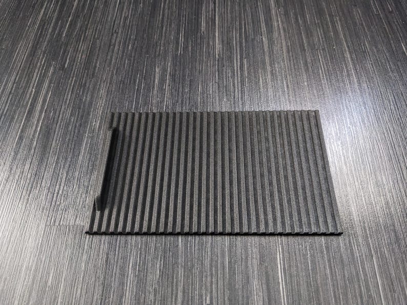 May include: A black, rectangular, ribbed, non-slip mat with a small cutout on the left side.