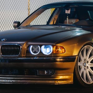 May include: A bronze-colored BMW sedan featuring a front grille with the BMW emblem, dual circular headlights with bright blue illumination, a lower front bumper with the text ALPINA, and large multi-spoke silver alloy wheels.