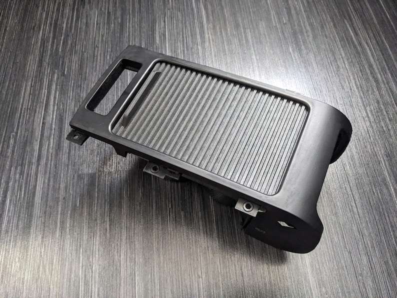 May include: Black plastic car part with a textured surface and a small handle. The part has a rectangular shape and is designed to fit into a car's interior.