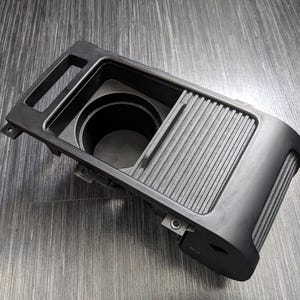 May include: Black plastic car cup holder with a sliding cover. The cup holder has a single cup slot and a sliding cover that reveals a storage compartment.