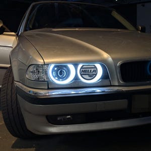 Op de afbeelding: Een zilverkleurige sedan met dubbele ronde koplampen voorzien van helderwitte LED-halo-ringen. De binnenste koplamp lens toont de zwarte tekst HELLA in een vetgedrukt lettertype. Het voertuig heeft een chromen sierstrip op de voorbumper en een zwarte grille.