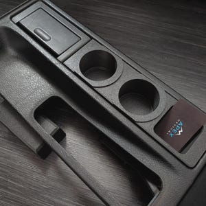 Può includere: Console automobilistica nera con superficie strutturata, dotata di un piccolo vano portaoggetti, due portabicchieri e un porta carte con il logo "Apex Designs". Progettata per interni di veicoli.