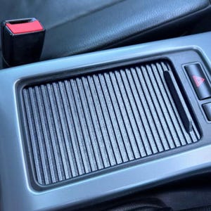 May include: A close-up of a car's interior, showing a gray, textured, rectangular panel with a black, ribbed insert. The panel has a small, red, triangular button on the right side.