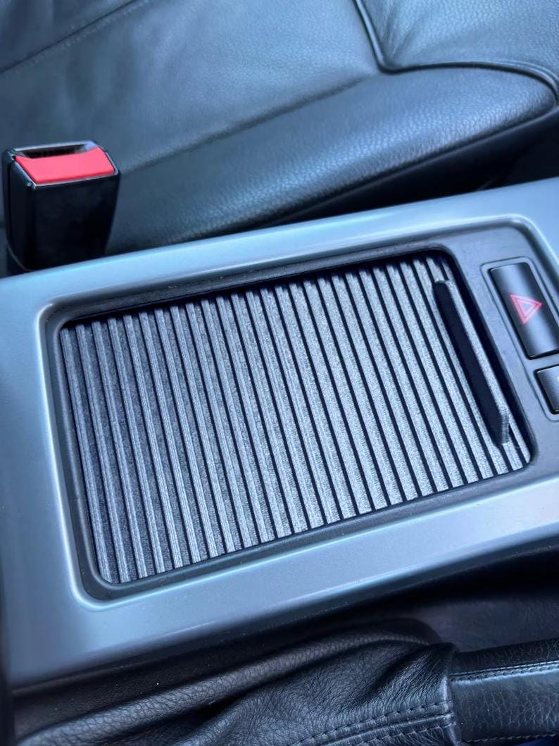 May include: A close-up of a car's interior, showing a gray, textured, rectangular panel with a black, ribbed insert. The panel has a small, red, triangular button on the right side.
