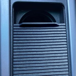 May include: A black plastic car cup holder with a sliding cover. The cup holder is located in the center console of a car. There are two buttons above the cup holder, one with a red triangle and the other with a picture of a car door.