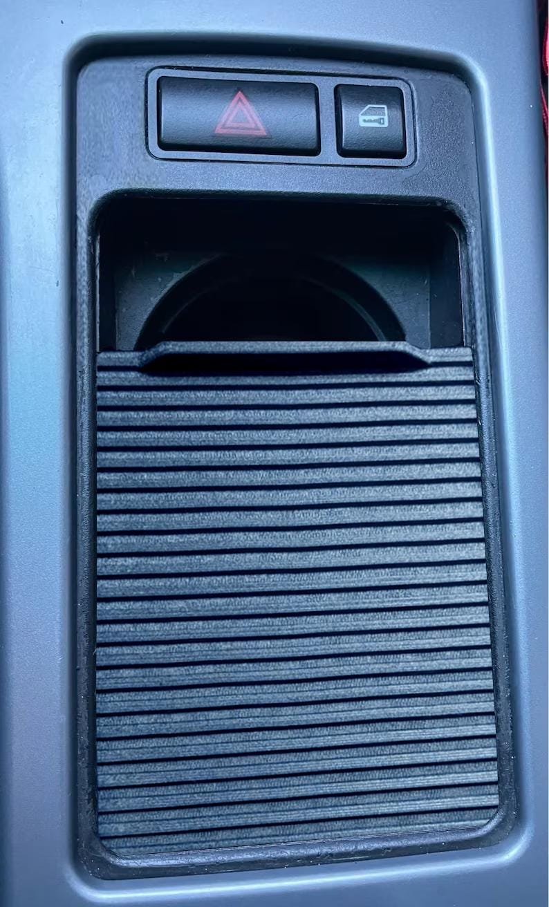 May include: A black plastic car cup holder with a sliding cover. The cup holder is located in the center console of a car. There are two buttons above the cup holder, one with a red triangle and the other with a picture of a car door.