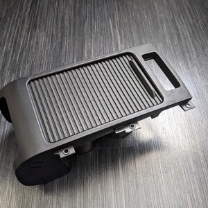 May include: Black plastic car part with a ribbed surface and a small handle. The part has several small holes and appears to be a storage compartment.