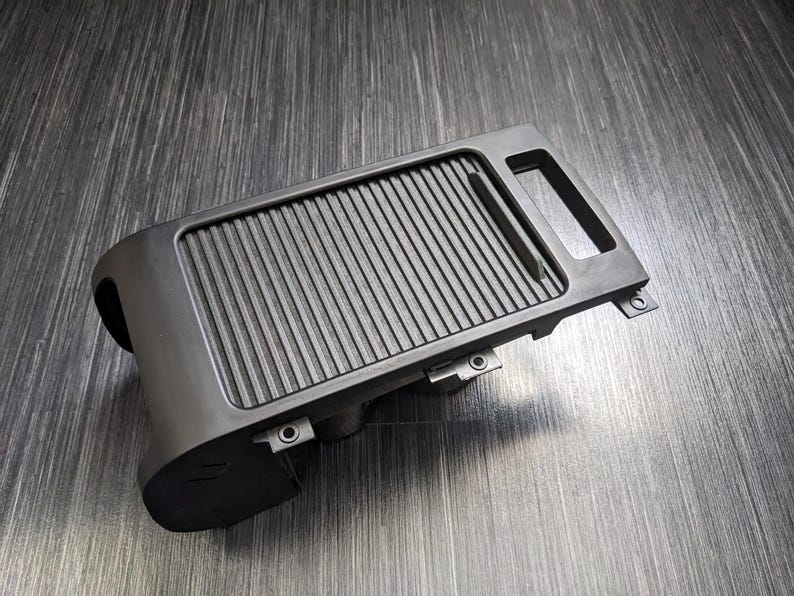May include: Black plastic car part with a ribbed surface and a small handle. The part has several small holes and appears to be a storage compartment.