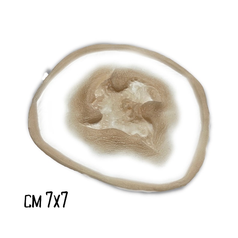 May include: A prosthetic wound with a light brown outer ring and a lighter, textured center. The text "CM 7x7" is printed in black at the bottom left corner. The wound is designed to simulate a skin injury.