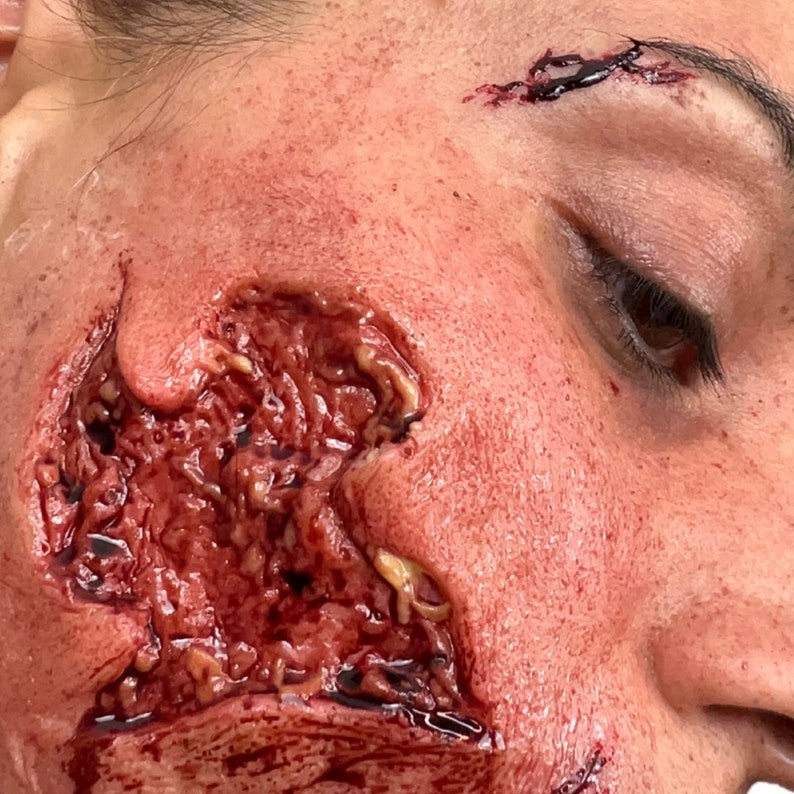 May include: A close-up of a face with a realistic wound prosthetic. The prosthetic features a deep, open wound with exposed tissue and blood. The skin around the wound is red and irritated, with additional cuts above the eye.
