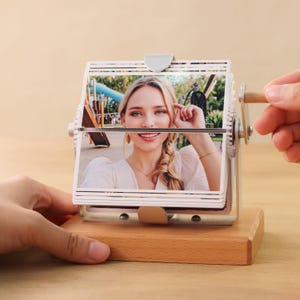 Puede incluir: Un expositor de fotos de madera con un mecanismo giratorio. El expositor presenta una foto de una mujer sonriendo. El expositor tiene una manivela en el lateral para girar las fotos.