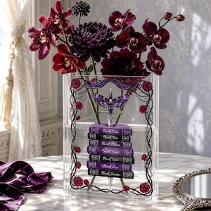 Può includere: Un vaso trasparente in acrilico pieno di fiori viola scuro, tra cui orchidee e dalie. Il vaso è decorato con un design gotico con una farfalla, una spada e una pila di libri con la scritta "Book One" fino a "Book Seven".
