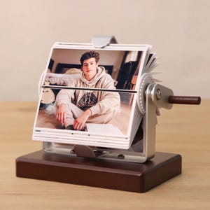 May include: A silver photo album with a wooden base. The album has a crank on the side to turn the pages. The album is filled with photos of a young man.