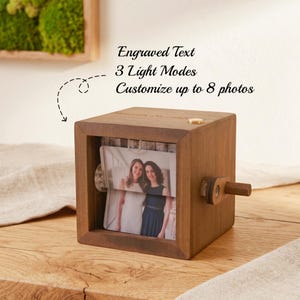 May include: A square wooden photo display box featuring a side crank handle, a top button, and an internal rotating mechanism. The text Engraved Text, 3 Light Modes, and Customize up to 8 photos is visible. A photograph of two people is displayed.