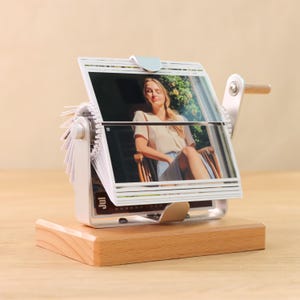 May include: A white photo album with a wooden base and a crank handle. The album is filled with photos of a woman in a white shirt and a skirt. The album is on a wooden table.