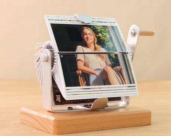 May include: A white photo album with a wooden base and a crank handle. The album is filled with photos of a woman in a white shirt and a skirt. The album is on a wooden table.
