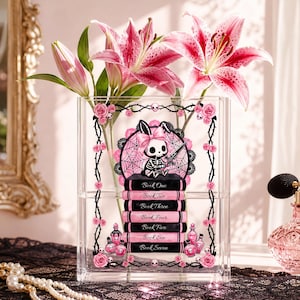 Può includere: Un vaso rettangolare in acrilico trasparente con la grafica di un coniglio scheletro con un fiocco rosa seduto su una pila di sette libri etichettati da Book One a Book Seven, circondato da una cornice di rovi neri con rose rosa.