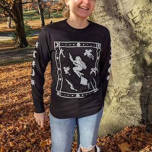 May include: A person wearing a black long-sleeved t-shirt with a white graphic of a witch flying on a broomstick surrounded by stars and moons. The shirt has a white border with a repeating pattern of stars and moons on the sleeves. The person is standing in a pile of autumn leaves.