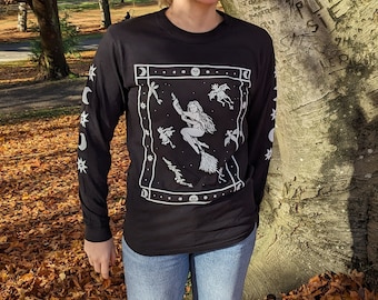 May include: A person wearing a black long-sleeved shirt with a white graphic of a witch flying on a broomstick surrounded by stars and moons. The shirt has a white border with a repeating pattern of stars and moons on the sleeves. The person is standing in a pile of autumn leaves.