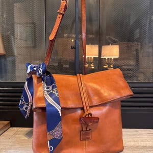 May include: A brown leather satchel bag with a flap closure and a long adjustable strap. A blue and white patterned scarf is tied to the strap. The bag has a wooden toggle closure and is displayed on a black stand.