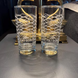 May include: Two clear shot glasses with white wave pattern designs and the "PATRON TEQUILA" logo. The glasses are cylindrical and sit on a dark surface. A gold-colored decorative object is in the background.