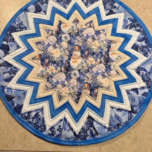 May include: A round, quilted placemat with a winter theme. The center features a floral pattern with snowmen. The design includes a starburst pattern in shades of blue, white, and tan, with a blue border. The placemat is approximately 14 inches in diameter.