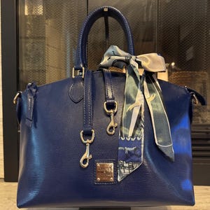 May include: A deep blue textured leather handbag featuring two top handles, a top zipper closure, and a silver-toned metal logo plate. A patterned silk scarf in shades of blue, white, and green is tied to one handle, and two silver-toned metal clips hang.