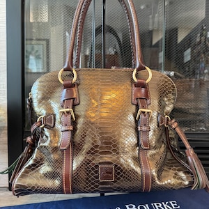 May include: A brown snakeskin-patterned handbag with brown leather accents, handles, and buckles. The bag has gold-colored rings and a tassel on each side. The brand name "DOONEY & BOURKE" is visible on a blue cloth.
