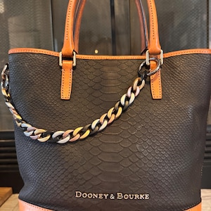 May include: Black tote bag with a snakeskin texture, tan leather trim, and handles. The bag features a multi-colored chain and the brand name "Dooney & Bourke" in silver lettering.