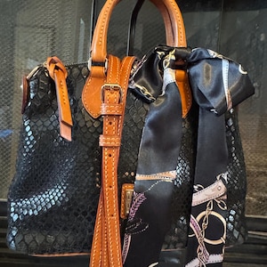 May include: Black handbag with a snakeskin texture, brown leather handles, and a matching shoulder strap. A black scarf with a chain and floral pattern is tied to the handle. The bag has a buckle closure.