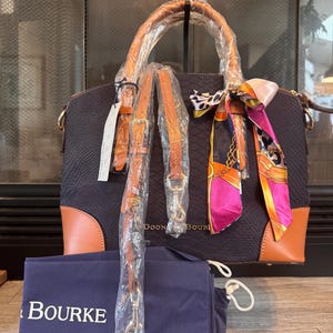 May include: A navy blue handbag with brown leather trim and handles. The bag has a textured pattern and comes with a detachable shoulder strap. A colorful scarf is tied to the handle. A navy blue dust bag with the word "BOURKE" is also included.
