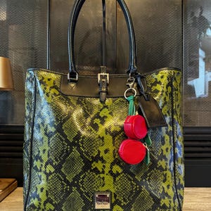 May include: A large tote bag with a snakeskin pattern in shades of green and black. The bag has black leather handles and trim, with a buckle closure and a small red coin purse attached. A small tag hangs from the handle.